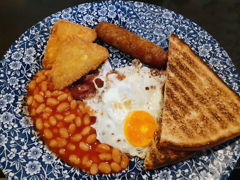 Nottingham, England, July 30, 2022. A typical English breakfast. The plate includes a fried egg, bacon, a pork sausage, white beans, two triangular pieces of grated and fried potatoes, two toast.