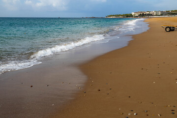 Calm turquoise sea with white surf line on yellow sandy beach under partly cloudy sky, showing distant resort umbrellas and palms during season's final warm days. Sorgun, Antalya, Turkey.

