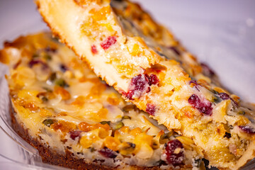 A thick slice of fruit and rhubarb cake topped with pumpkin seeds, dried cranberries and apricots, removed from a round baked cake. Rich texture and visible moist layers.