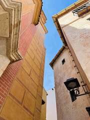 A bottom-up view of downtown buildings, painted brick patterns on a plaster wall, a decorative metal street lamp on the wall, salmon and terracotta tones. Malaga, Spain, 2022.