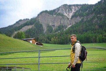 Hiker standing by a metal fence in a green mountain valley with forested hills and rocky cliffs in the background.