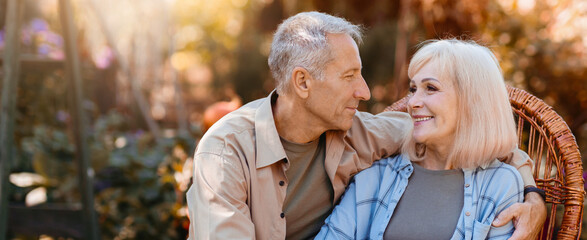 A senior couple sits closely on a comfortable chair in a vibrant garden. They share loving smiles and gaze into each other's eyes, enjoying a peaceful moment in the sunlight.