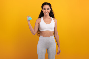 A cheerful woman in sportswear stands confidently in front of an orange background. She is holding a dumbbell in one hand, showcasing her fit lifestyle and enthusiasm for fitness activities.
