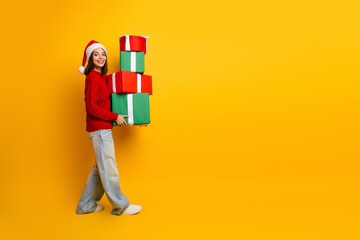 Funny Christmas shopper girl carries colorful gifts against bright yellow wall