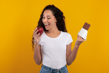 Young woman with curly hair smiles while holding a chocolate bar in one hand and a red apple in the other. She is engaged in making a joyful food choice, set against a vibrant yellow-orange wall.