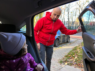 A parent leans toward the open car door to check on a child seated inside. The moment captures everyday parenting, attention to child safety, and preparing a child for a car ride