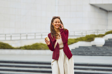 Fototapeta premium A stylish woman wearing a burgundy blazer and beige dress chats on her phone while holding a coffee cup in an urban outdoor setting. Bright, cheerful mood with a casual business vibe.