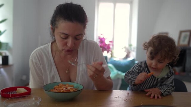 Mother eating lunch beside toddler who nibbles food with quiet curiosity, creating calm shared moment at kitchen table filled with closeness and gentle family presence