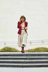 Obraz premium Stylish businesswoman in burgundy jacket walking on the street and talking on phone with sustainable coffee cup to go. 