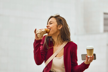Businesswoman in a rush taking a bite of a croissant outdoors, rushing to office while holding smart phone and coffee to go. Casual lifestyle moment, street food vibes, urban breakfast scene.