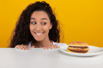 A joyful lady looks eagerly at a tempting burger on a plate, peeking over a table in a bright yellow-orange studio. She symbolizes cravings and the allure of cheat meals.