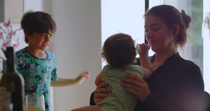 Mother holding toddler while older child plays nearby, sharing warm affectionate moments together in bright home as siblings interact with joy and gentle family connection