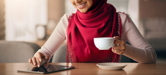 A young woman wearing a red scarf sits at a table in a warm cafe, sipping coffee from a white cup. She is engaged with her tablet, smiling softly and enjoying her time.