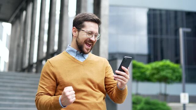 Happy excited businessman received great news on mobile phone standing on the street near modern office building. Smiling shocked surprised worker in glasses reads positive good message on smartphone