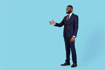 Cooperation Concept. Smiling african american businessman giving hand for handshake aside while standing over blue studio background, black male entrepreneur in suit offering partnership, copy space
