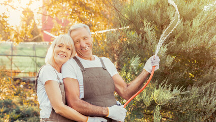 A happy couple stands close while watering plants in a vibrant garden. The warm sun shines on them as they enjoy their time together, surrounded by greenery and autumn colors.