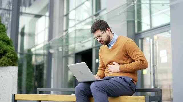 Businessman feels stomach pain while working on a laptop sitting on a bench on street near business office building. Sick man has heartburn, gastritis, bloating or poisoning. Male suffers from spasms