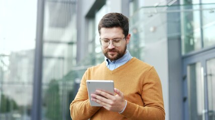 Confident businessman is using digital tablet while standing on street near an office building. Smiling entrepreneur in casual clothes checking email messages, chatting online, texting, browsing web - Powered by Adobe
