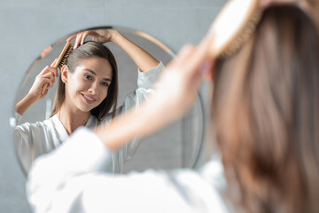 Naklejka premium A young attractive lady stands in front of a mirror, carefully brushing her hair. She smiles at her reflection while embracing a self-care moment in her bathroom.