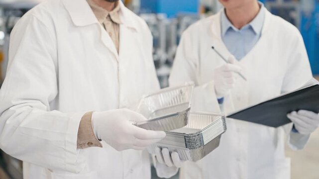 Close up of lab workers in protective gloves and coats holding aluminum trays and clipboard. Inspecting product quality and discussing specifications at food packaging factory. Industrial environment.