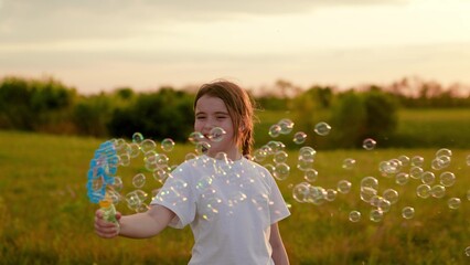 Happy child girl having fun on lawn blowing soap bubbles. Smiling girl child playing with soap bubbles in park in summer. Child girl rejoices blowing soap bubbles on playground, flying soap bubbles