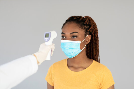 A young black female patient wears a face mask and checks her body temperature with an electronic thermometer. This occurs in a grey studio background as part of the Covid-19 vaccination process. - Powered by Adobe