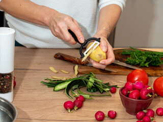 Woman peeling a potato with a vegetable peeler while preparing fresh vegetables on the countertop. Healthy home cooking.