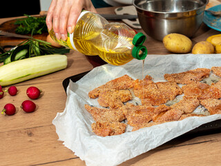 Pouring oil over breaded meat pieces placed on baking paper. Home cooking and dinner preparation.