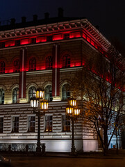 Obraz premium A historic building in Riga illuminated at night with striking red and warm white lights, accented by ornate street lamps and autumn trees, creating a dramatic and atmospheric city scene.