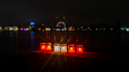 Row of memorial candles glowing at night with a blurred city skyline in the background. Warm red and white candle lights create a soft, atmospheric scene with colorful bokeh and peaceful mood.