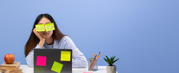 A student sits at a desk covered in study materials, looking bored with sticky notes on her face....