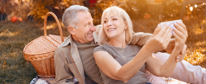 A happy senior couple relaxes on the grass during a sunny day in the park. They share laughter while taking selfies, with a picnic basket beside them full of treats. - Powered by Adobe