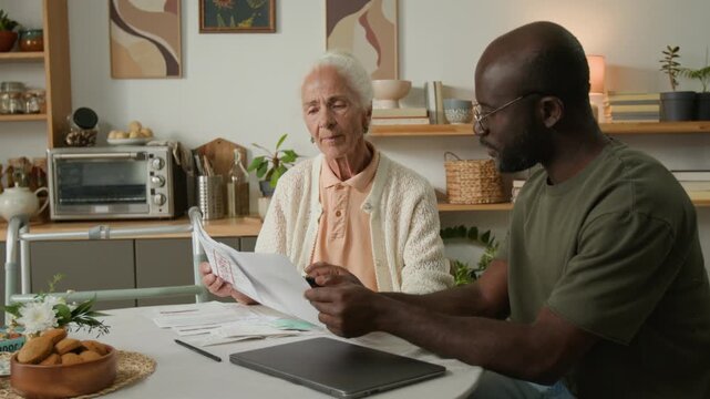 Medium shot of white haired senior woman doing tax payments and learning about home finances with Black man helping with paying bills while sitting at table together and talking