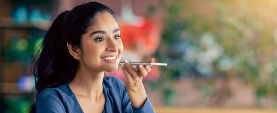 A young woman smiles while speaking into her smartphone, likely using a voice assistant. The setting is a cozy cafe filled with greenery and soft lighting, creating a welcoming atmosphere. - Powered by Adobe