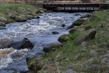 Rough Stream Flowing Over Mossy Rocks Toward Wooden Bridge in Natural Riverbed Landscape