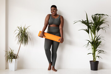 Young african american woman stands by a white wall smiling while holding a rolled fitness mat. She...
