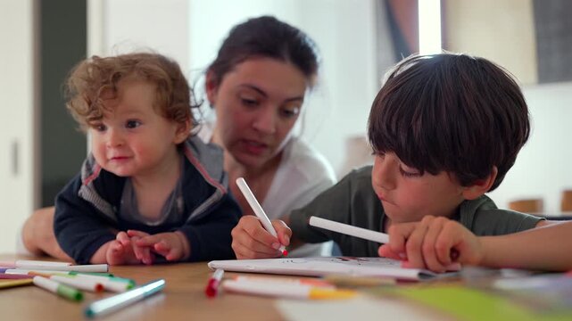 Baby sits beside mother watching older brother draw with markers at table creating cozy household moment of gentle attention curiosity shared focus and early creative engagement among siblings