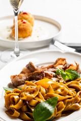 A rich restaurant dish of tagliatelle pasta with a slow-cooked meat ragù, an egg yolk, basil leaves, and a glass of sparkling wine, with an appetizer in the background