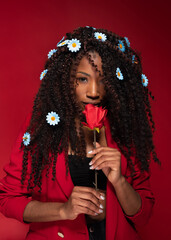African american woman with curly long hair smelling red rose with daisies in hair on red background