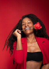 Studio shot of cheerful Hispanic woman smiling and holding rose in mouth against red background