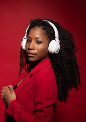 Hispanic woman with curly hair in red blazer wearing headphones listening music on red background