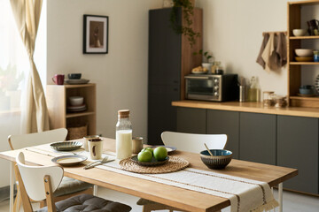 Modern kitchen interior featuring wooden dining table set with breakfast items including milk, apples, bowls and cups, sunlight streaming through window, cozy home atmosphere