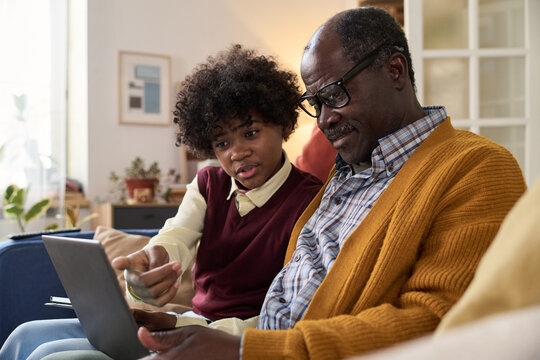 Black senior man sitting with Black teenager using digital tablet together, teenager pointing at screen while explaining something, both focused on device in home setting