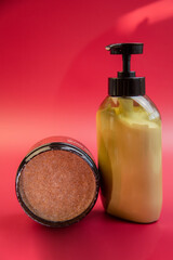 Beauty products showcased on a bright red backdrop, including an exfoliating scrub jar and a moisturizing lotion pump bottle, ideal for skincare routines