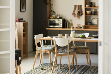 Modern kitchen interior featuring wooden dining table set with plates, cups, milk bottle and fruit, striped rug on floor, open shelving with kitchenware in background, no people visible