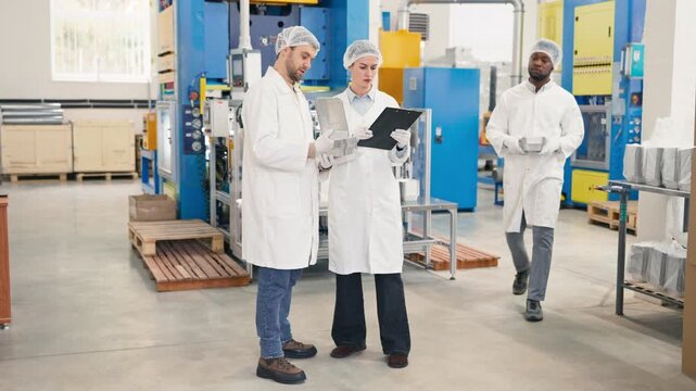 Three lab colleagues wearing protective coats and hairnets checking aluminum trays and discussing production quality. Team conducting inspection at food packaging factory. Industrial machinery.