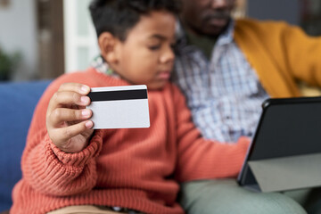 Black child holding credit card while sitting next to Black man using digital tablet, both focusing on online activity, close up on hand with card, family spending time together