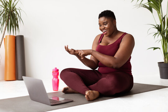 A cheerful plump black woman sits on a mat in a fitness studio, engaging with her students through a laptop during an online yoga class. A water bottle rests nearby. - Powered by Adobe