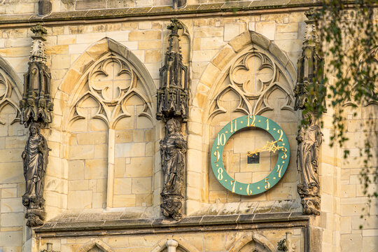 A wall element of a Gothic building, on which numerous figures made of stone in the Gothic style are stuck. There is also a gold-colored clock on the wall, which shows the time 10 minutes past 3