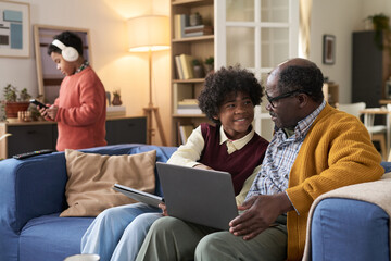 Black senior man sitting on sofa using laptop with Black teenage boy while Black child standing in background using digital tablet in living room, family spending time together